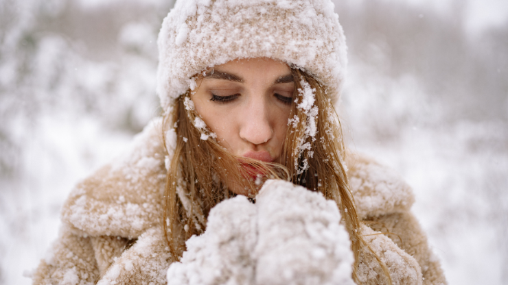 Frau im Winter mit Mütze und Handschuhen, die in kalter, verschneiter Umgebung in ihre Hände pustet. Symbolbild für Hautschutz und Gesichtspflege bei Frost und Kälte.